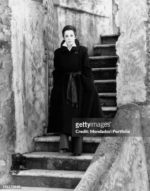 The Italian actress and singer Daniela Goggi, wearing a dark coat, is standing with absorbed expression on the stairs leading to a dock along the...