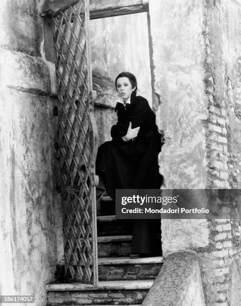 The Italian actress and singer Daniela Goggi, wearing a dark coat, is standing upright with a foot leaning on a gate, with crossed arms and absorbed...