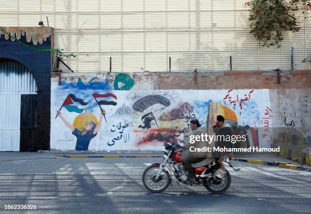 Yemeni men riding a bike drive next to murals depicting solidarity with Palestinians painted on the wall of the closed Saudi Arabia embassy on...