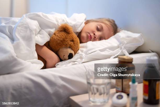 a sick girl sleeps under a blanket with a teddy bear in her arms after taking medications on the bedside table. - bedside table kid asleep stock pictures, royalty-free photos & images