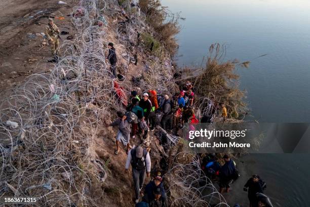 Texas National guardsman watches as migrants pick their way through razor wire after crossing the Rio Grande into the United States on December 17,...