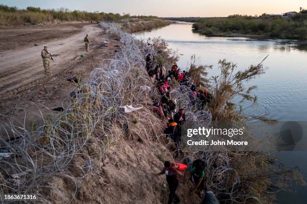Texas National guardsmen watch as migrants pick their way through razor wire after crossing the Rio Grande into the United States on December 17,...