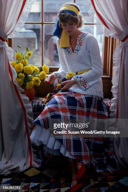 Portrait of model, dressed in a crocheted white sweater with floral applique and a plaid skirt with white petticoat, as she sits in a window seat,...