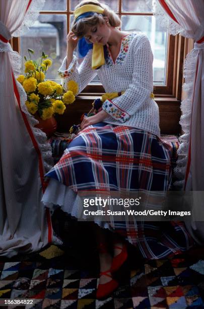 Portrait of model, dressed in a crocheted white sweater with floral applique and a plaid skirt with white petticoat, as she sits in a window seat,...
