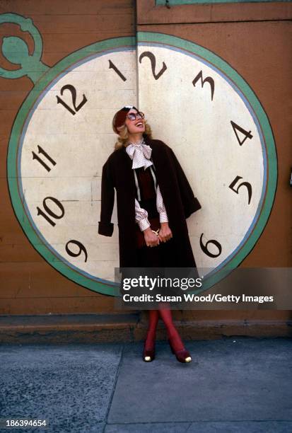 Portrait of a model dressed in a striped sweater vest over a cream-colored, bow-tie blouse, a brown skirt, and red tights with a brown coat draped...