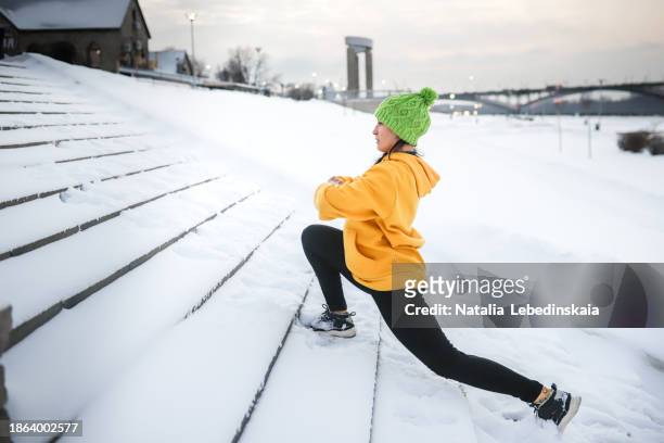 snowy urban exercise: woman in yellow sweatshirt and knit hat stretching on snow-covered stairs - sports training drill stock-fotos und bilder