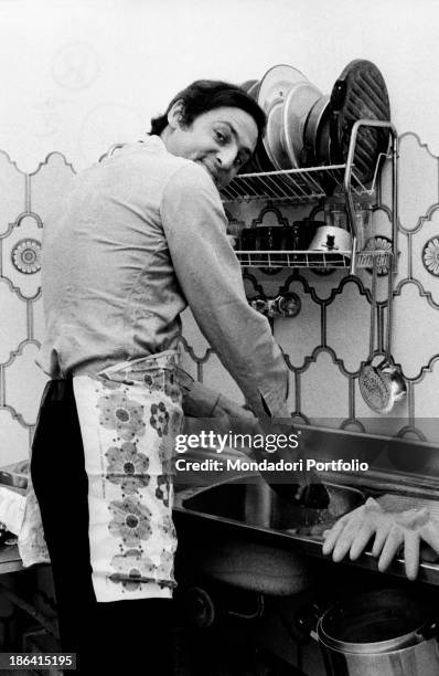 Pugliese musician and broadcast presenter Renzo Arbore washes the dishes in the kitchen sink wearing an apron like a perfect housewife, and smiles at...