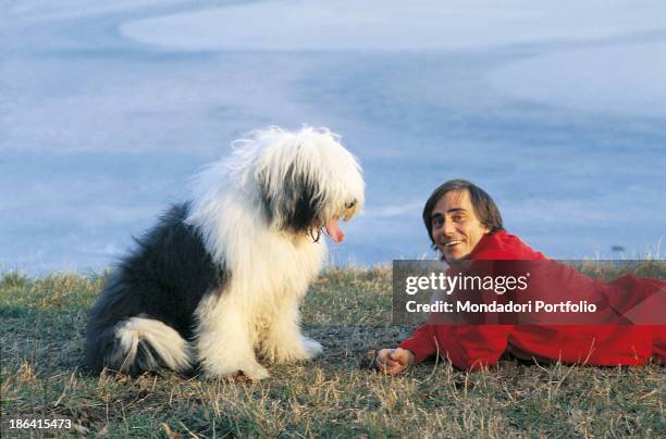 Italian singer-songwriter and writer Roberto Vecchioni smiling lying on a meadow beside a Bobtail dog. 1981.