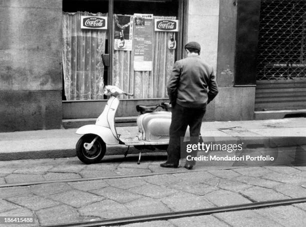 Man stopping to observe a Vespa parked outside a bar. Milan, April 1963.