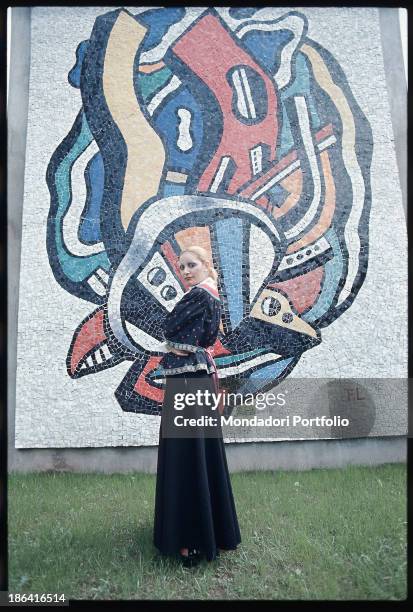 Italian singer Mia Martini posing with her arms crossed in the park of the Enzo Pagani Museum next to a mosaic made by the French artist Fernand...