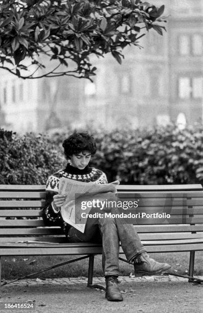 Young man reading a newspaper sitting on a bench. Italy, 1970s.