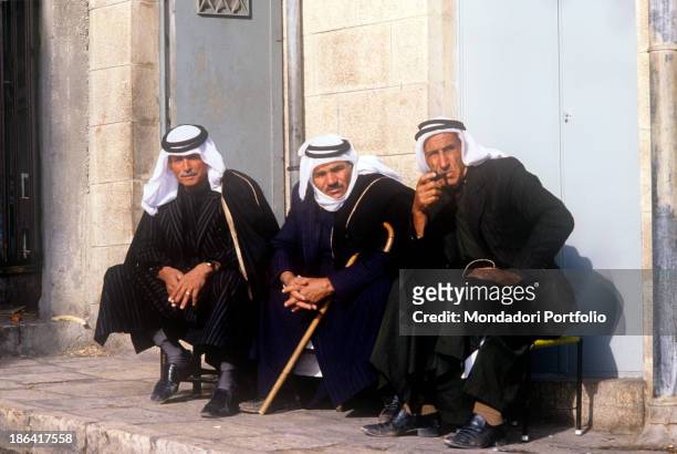 Three Palestinian men in traditional clothes sitting and smoking on the roadside. Palestinian Territories, January 1984.