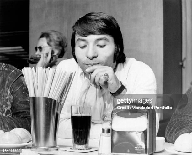 The drummer Alfio Cantarella, member of the Italian music band Equipe 84, is enjoying his drink with a straw seated at a café; in front of him a...