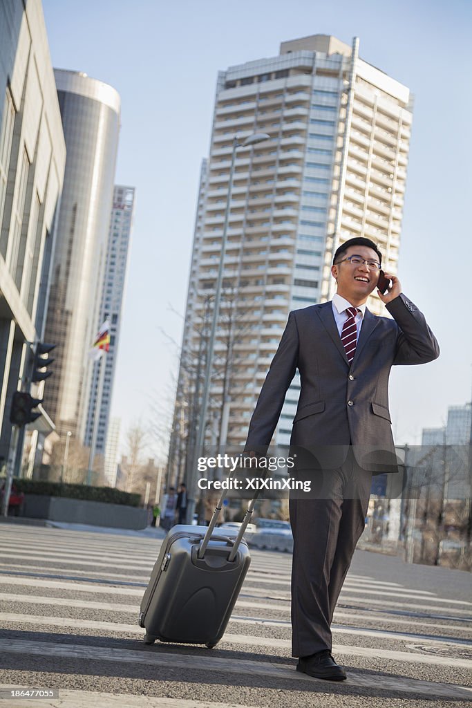 Smiling young Businessman walking down the street with luggage and on the phone