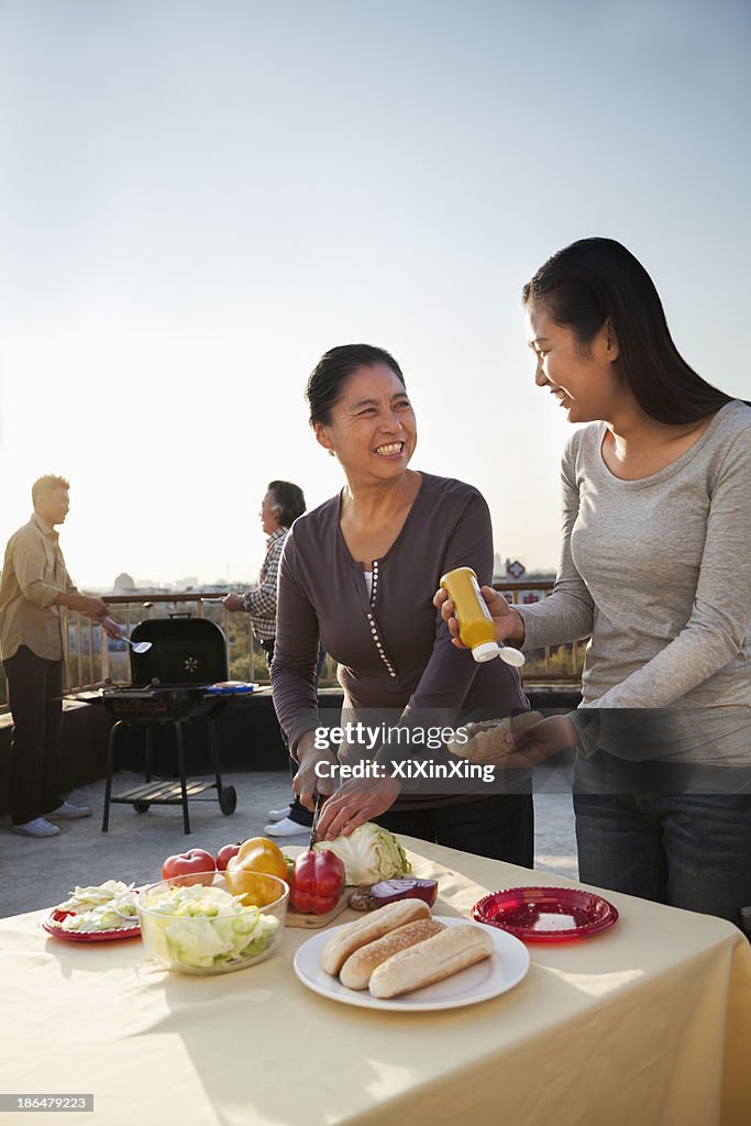 Mother and daughter preparing hot dogs for barbeque, father and son preparing sausages on barbeque
