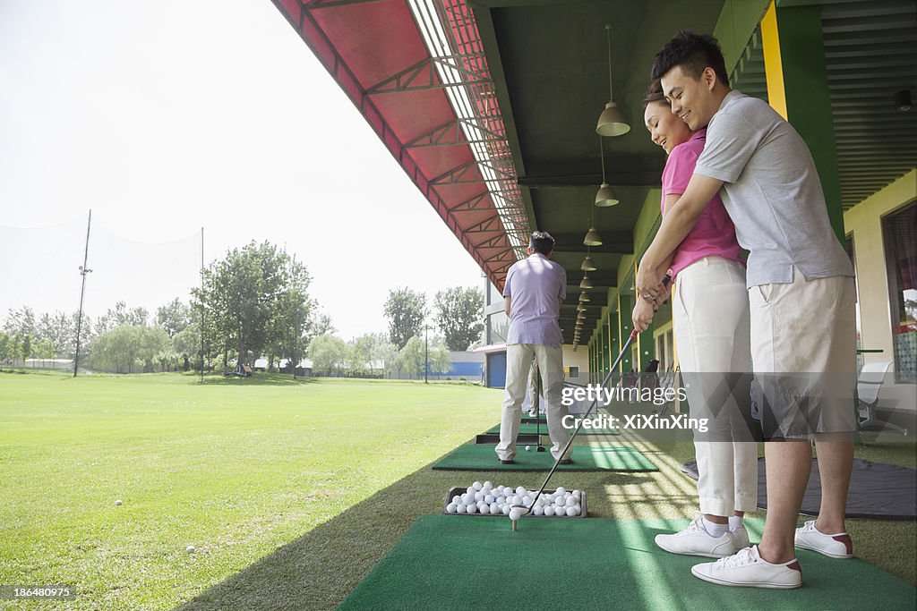 Young man teaching his girlfriend how to hit golf balls, arm around, side view