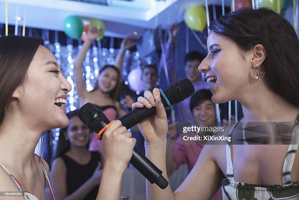 Two friends holding microphones and singing together at karaoke, face to face, friends in the background