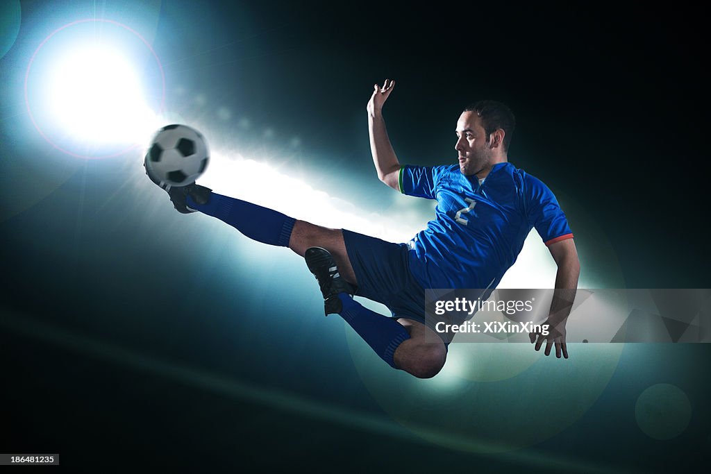 Soccer player in mid air kicking the soccer ball, stadium lights at night in background
