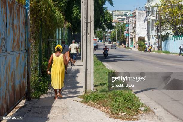 Havana, Cuba, A Cuban woman and other people walk in city sidewalk showing the lifestyle of real people. There is a large metal door and fence.