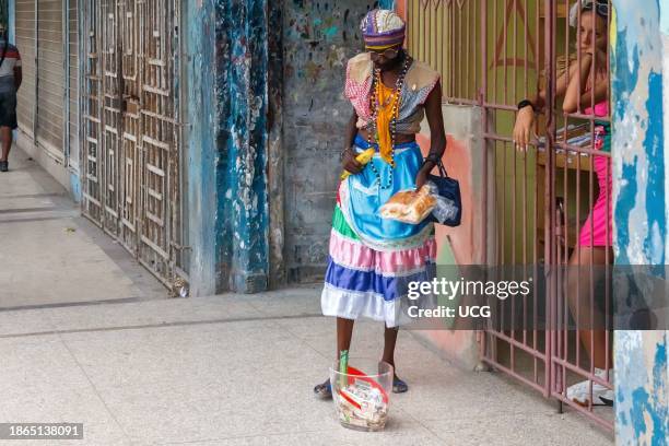 Havana, Cuba, A Cuban woman wearing Santeria clothing, necklaces and food in her hands begs for money a porch. Other people look at the scene from...