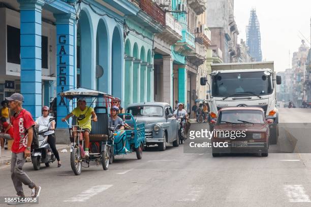 Havana, Cuba, People and traffic in a city street. A Russian Lada car is in front of a truck. A vintage car, tricycle, and a bicitaxi drive in the...