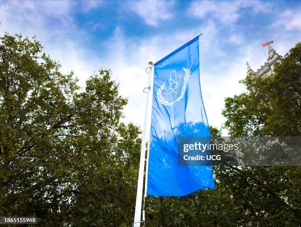 The logo of the International Atomic Energy Agency on a blue flag flying next to trees outside the Queen Elizabeth II Centre in London, UK.