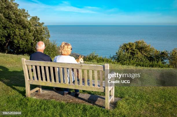 An elderly retired couple and their pet dog sit on a public bench and look out over the English Channel at Folkestone, Kent, UK.