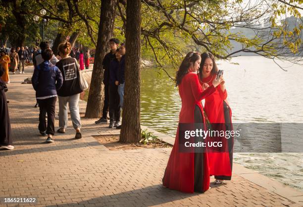 Two Vietnamese women in vivid red traditional ao dai stop to take a selfie next to Hoan Kiem Lake at Tet in Hanoi, Vietnam.