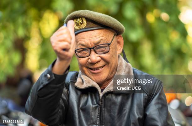 An elderly Vietnamese man wearing an olive green army-style beret and leather jacket gives a thumbs up in Hanoi, Vietnam.