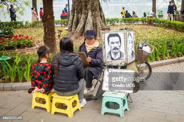 Male Vietnamese street artist draws a portrait of a mother and daughter in winter next to Hoan Kiem lake in Hanoi, Vietnam.