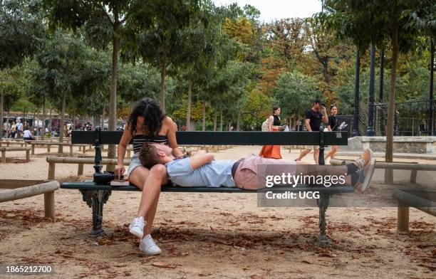 Young man lies on a bench resting his head on his girlfriend's lap as she looks at a mobile phone in Jardin du Luxembourg, Paris, France.