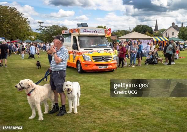 Man with holding two white dogs on leads eats an ice cream next to an ice cream van at Wisborough Green Village Fete, West Sussex, UK.