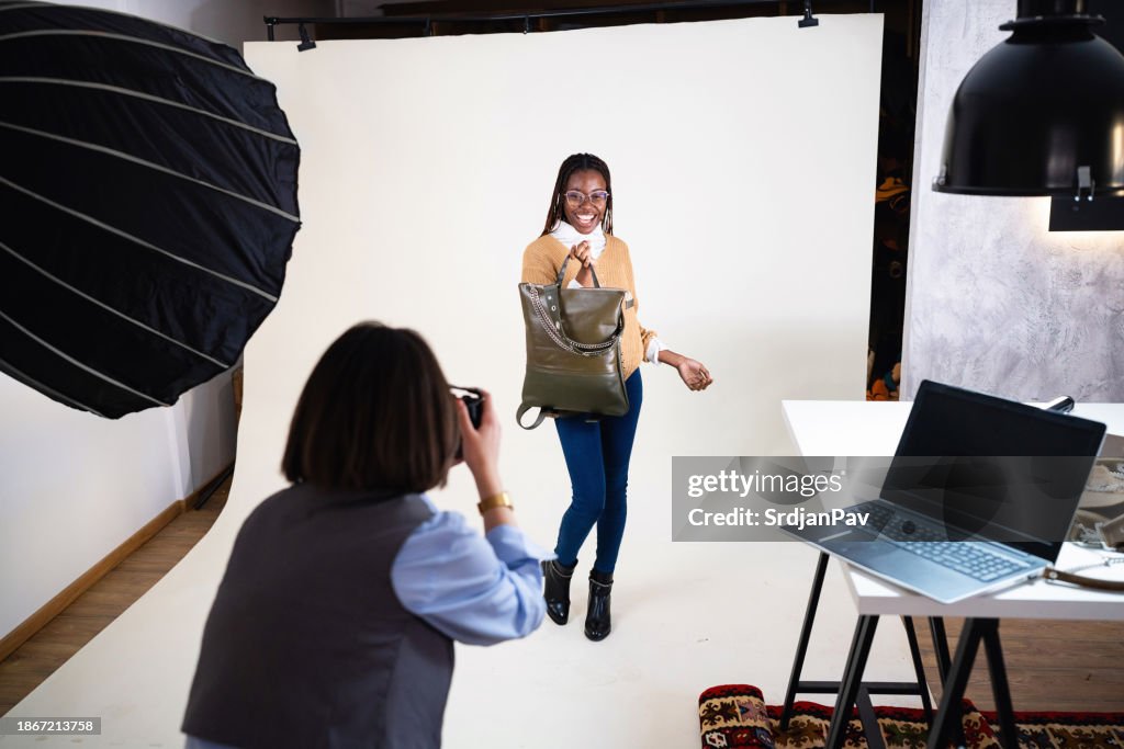Caucasian female photographer, taking photos of the female fashion model Black ethnicity, while posing with an leather bag