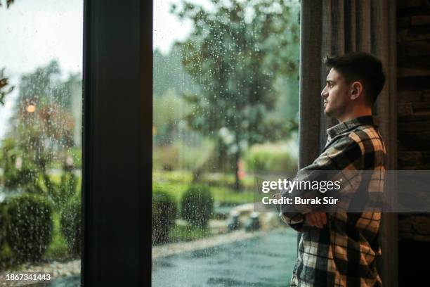 hombre joven mirando por la ventana en un clima lluvioso, meses de invierno, clima frío - mirar-por-la-ventana fotografías e imágenes de stock