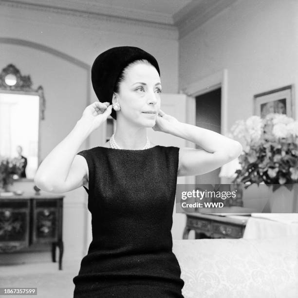 Outtake; Gloria Guinness adjusting her Balenciaga hat in her suite in the Waldorf Towers wearing a black wool Givenchy jacket bordered in black...