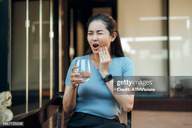 asian  adult woman hypersensitive teeth pain after drinking cold water with ice while sitting in the yard at home - human teeth stock pictures, royalty-free photos & images