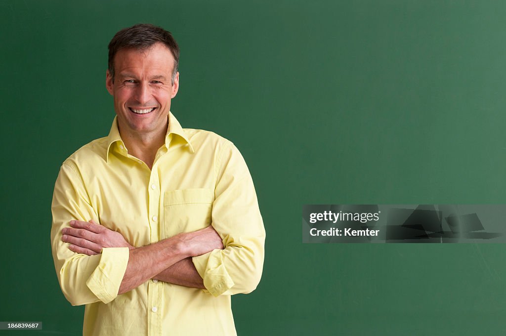 A smiling teacher with arms crossed in front of chalkboard