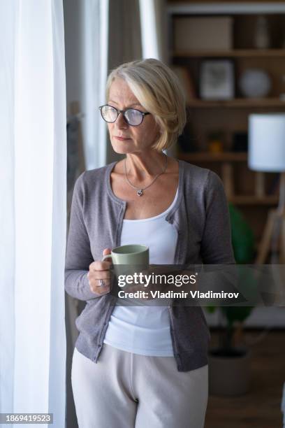 reife frau schaut aus dem fenster - winterdepression stock-fotos und bilder