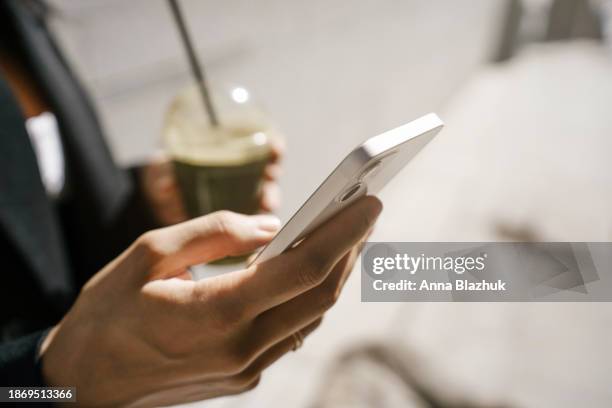 close-up of female hand holding phone in one hand and smoothie in cup in other hand standing outdoors on street in bright sunlight - smoothie close up textfreiraum stock-fotos und bilder