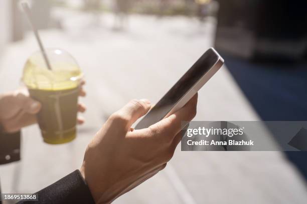 close-up of female hand holding phone in one hand and smoothie in cup in other hand standing outdoors on street in bright sunlight - smoothie close up textfreiraum stock-fotos und bilder