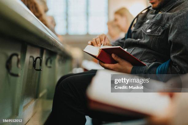 midsection of young man flipping page of hymnbook while sitting at protestant church - banco de igreja imagens e fotografias de stock