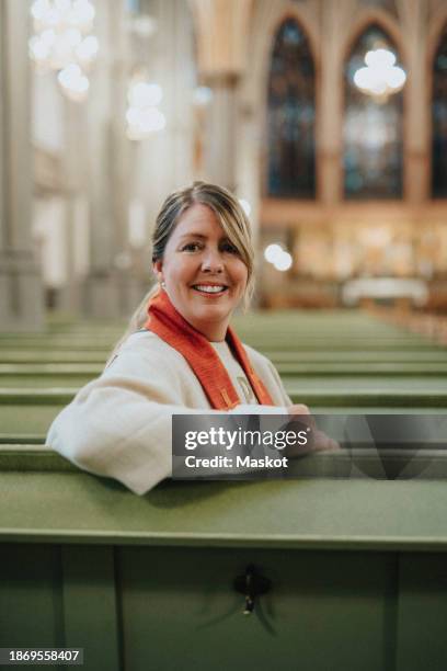 portrait of smiling female priest sitting on pew at protestant church - priest stock pictures, royalty-free photos & images
