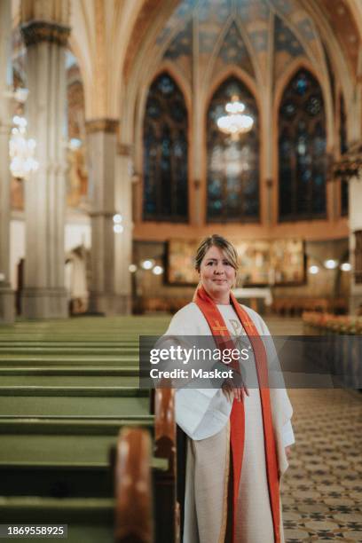 portrait of smiling female priest leaning on pew while standing at protestant church - protestantism stock pictures, royalty-free photos & images