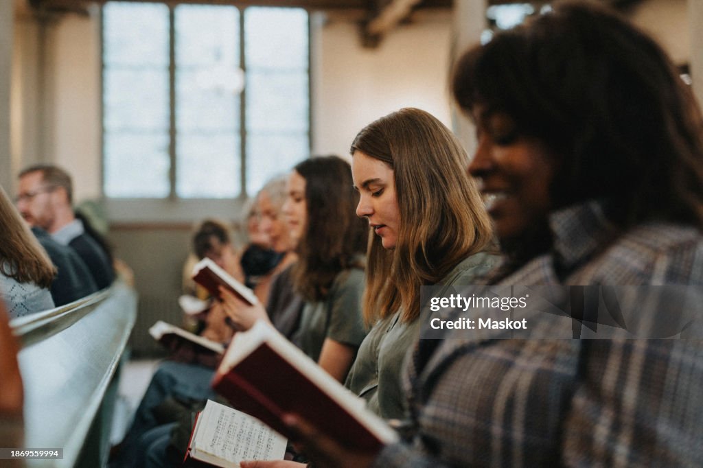 Young woman reading hymnbook while sitting with protestants at church