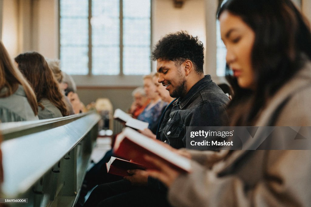 Smiling young man reading hymnbook while sitting with protestants at church