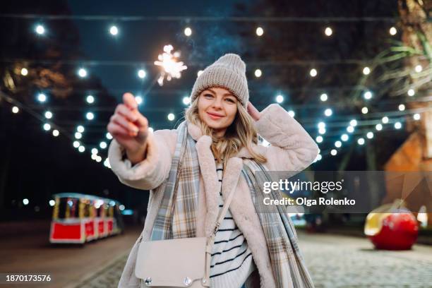 holidays are coming with sparklers and christmas lights. smiling woman in fur coat, lighting a sparkler on night streets - bengalischer feuer stock-fotos und bilder