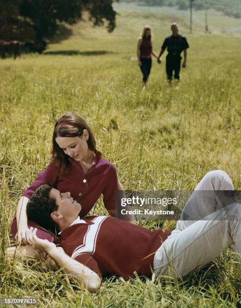 Young couple sitting on the grass, the man wearing a red-and-white sweater and grey trousers has his head in the lap of the woman, who wears a red...