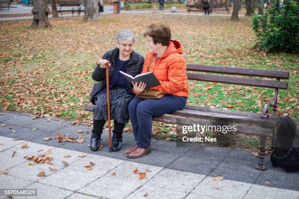 the color of autumn leaves, two women sitting on a park bench - park bench stock pictures, royalty-free photos & images