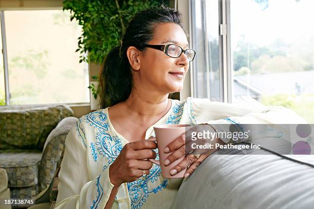mixed race woman having cup of coffee on sofa - adult woman cup tea stockfoto's en -beelden