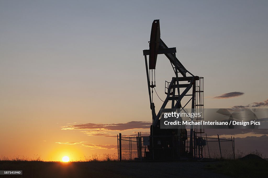 Silhouette Of Pump Jack With The Orange Glow Of The Sun Rising On The Horizon